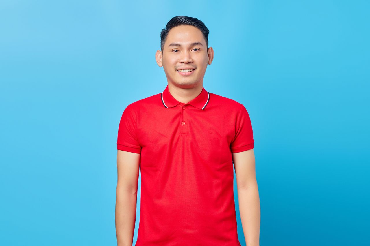 Cheerful young adult man wearing a red polo shirt, posing against a bright blue backdrop.