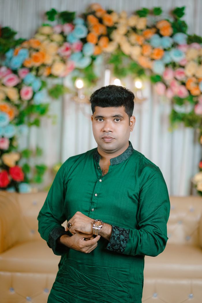 A groom in traditional attire in front of a floral backdrop at a Bangladeshi wedding.